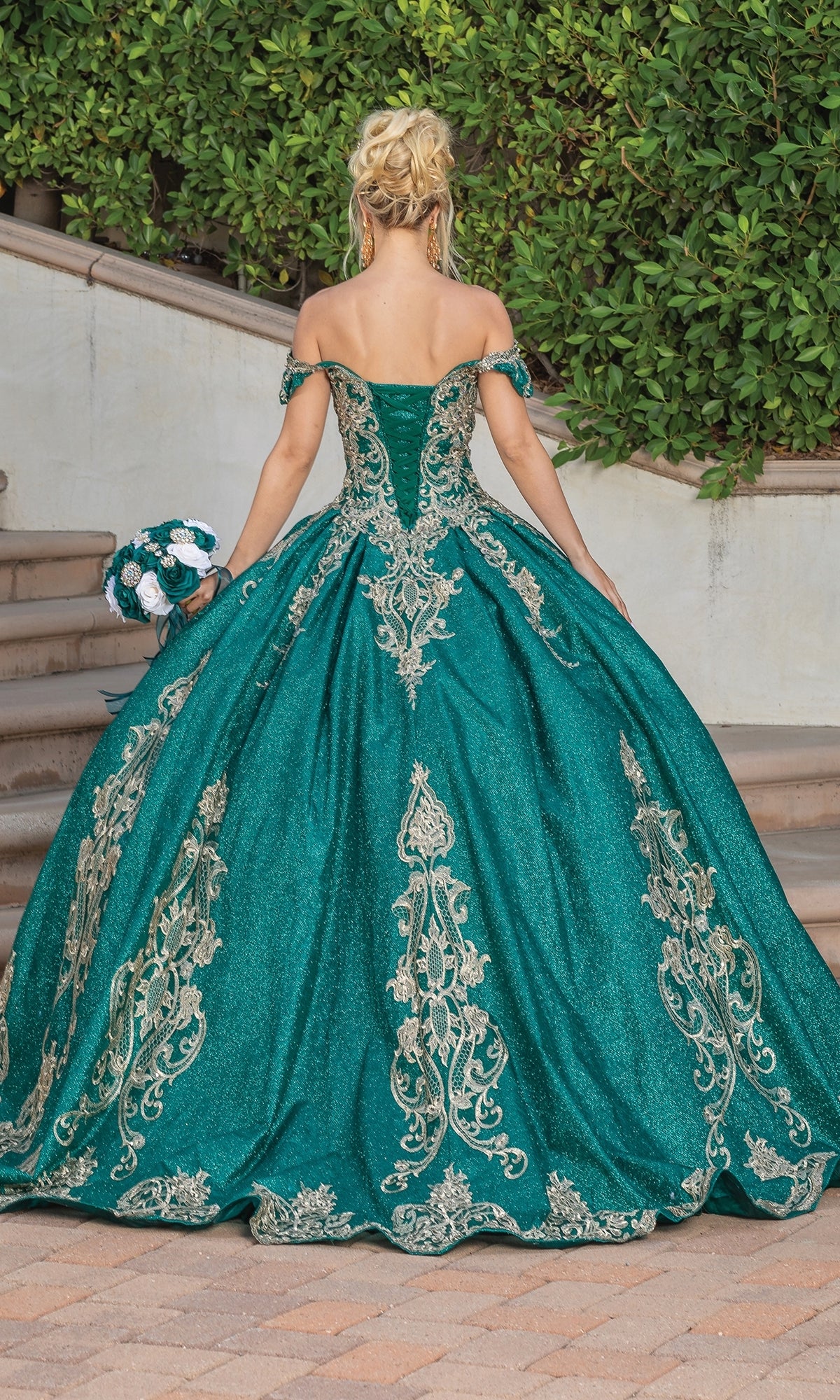 A woman stands outdoors, facing away in the Dancing Queen Quinceanera Dress 1769—a hunter green ball gown with gold lace embroidery, off-the-shoulder style—holding a matching bouquet.